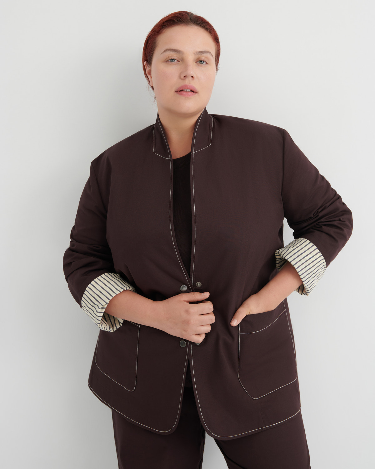 A woman modeling a dark brown jacket with contrasting light topstitching and striped button-up cuffs, paired with matching dark brown trousers against a light gray studio background.
