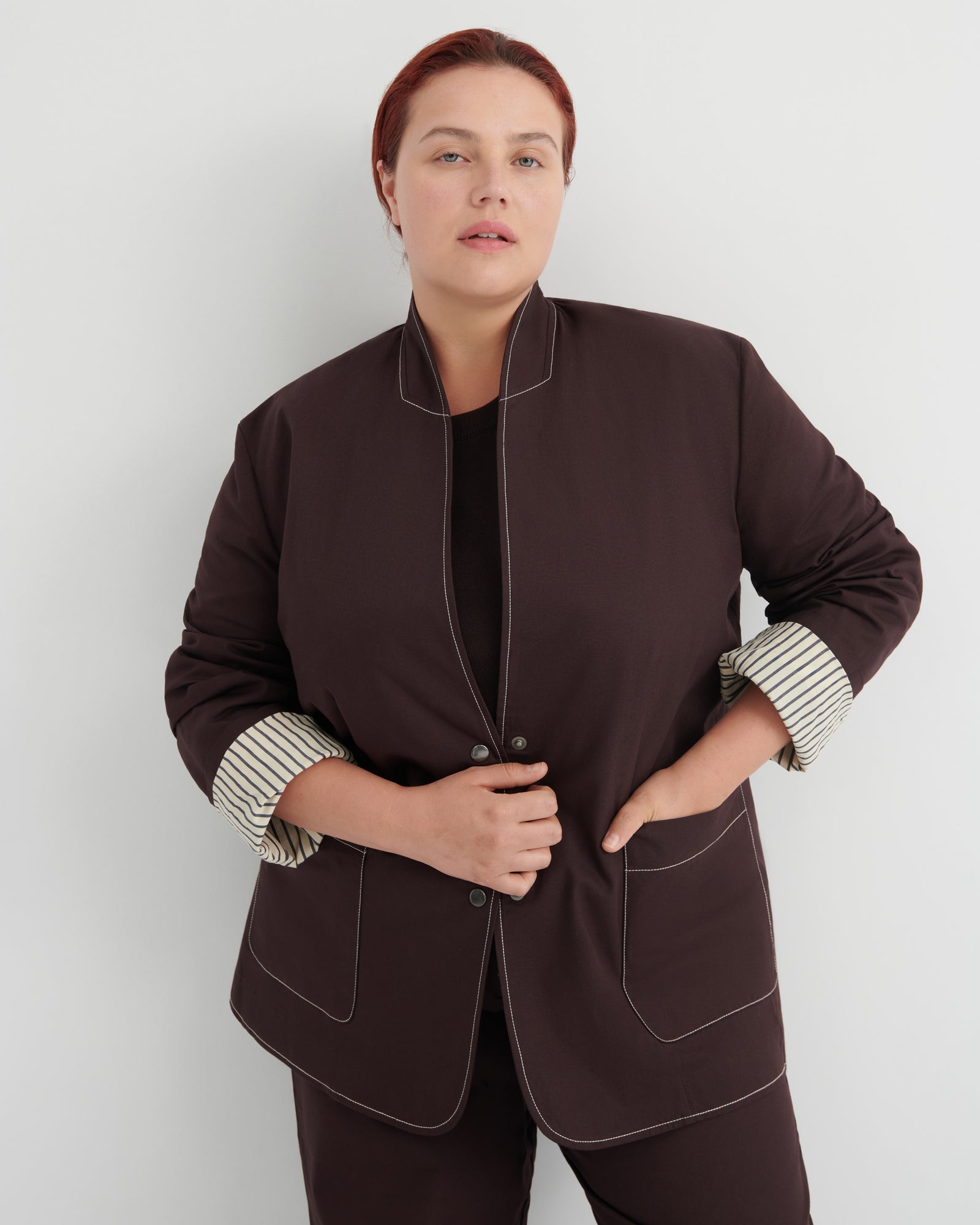 A woman modeling a dark brown jacket with contrasting light topstitching and striped button-up cuffs, paired with matching dark brown trousers against a light gray studio background.
