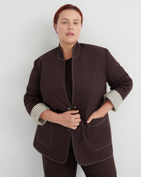 A woman modeling a dark brown jacket with contrasting light topstitching and striped button-up cuffs, paired with matching dark brown trousers against a light gray studio background.