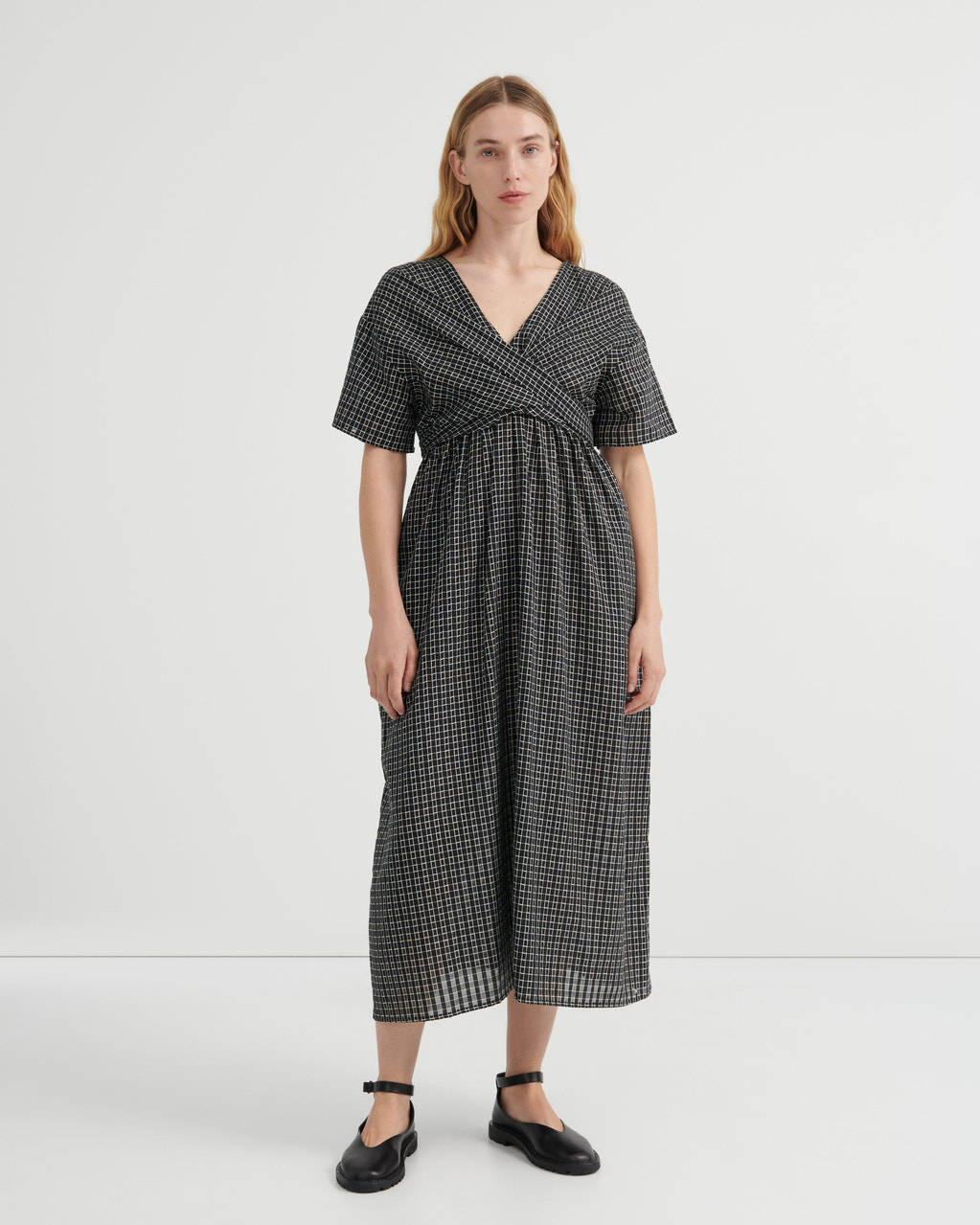 A woman modeling a short-sleeve wrap-style midi dress with a black and white micro-check pattern, standing against a plain light gray studio background.