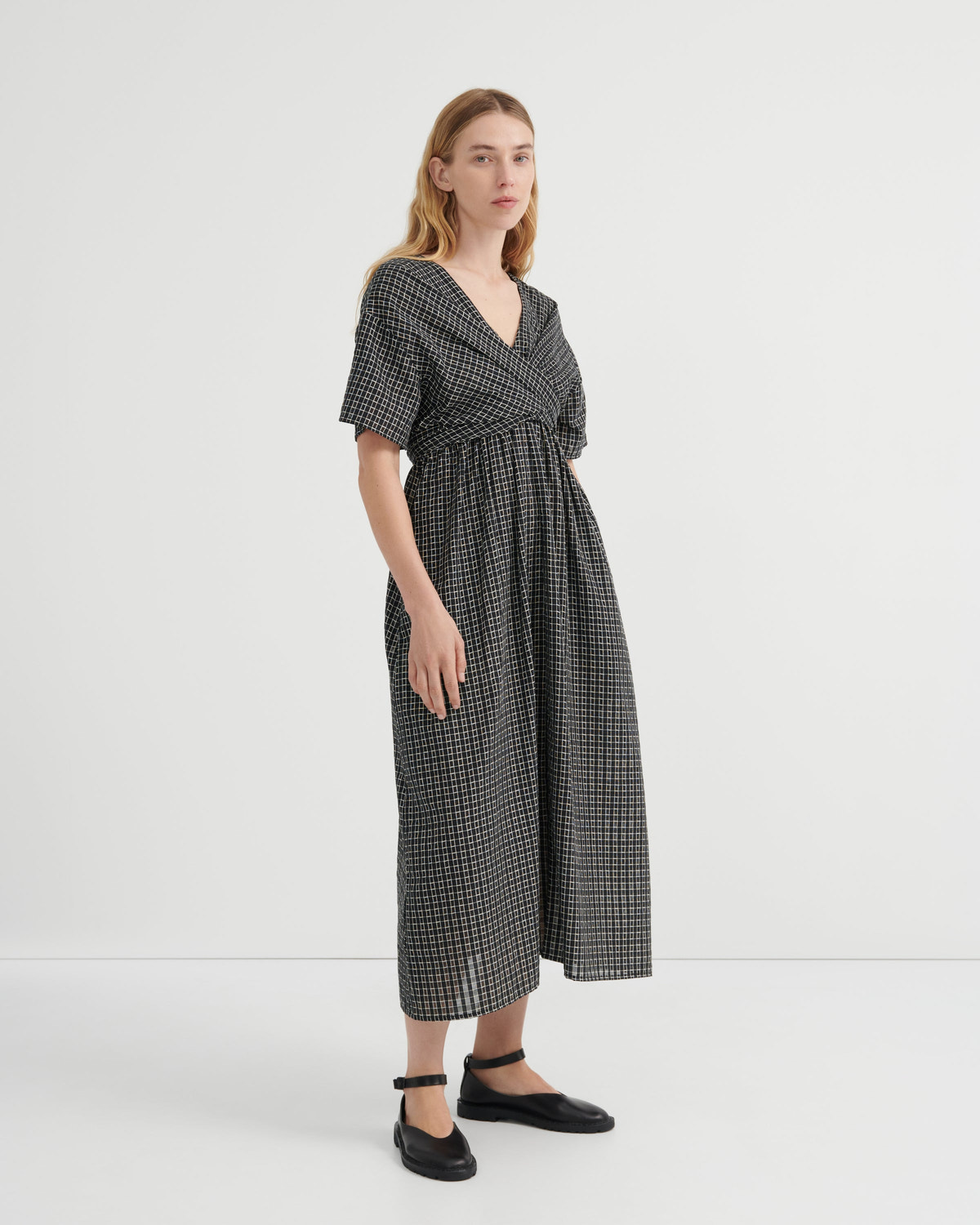 A woman modeling a black and white micro-checkered midi wrap dress with short flutter sleeves and a tie waist, paired with black Mary Jane flats, against a plain white studio background.