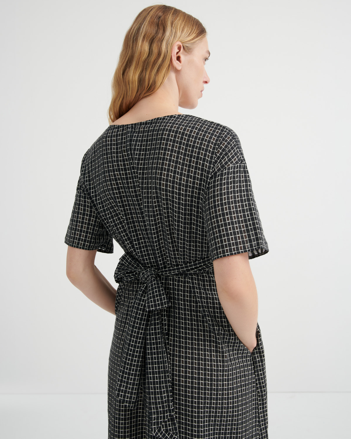 A woman shown from the back wearing a short-sleeve black dress with a white windowpane plaid pattern and a matching waist tie, set against a plain light gray studio background.
