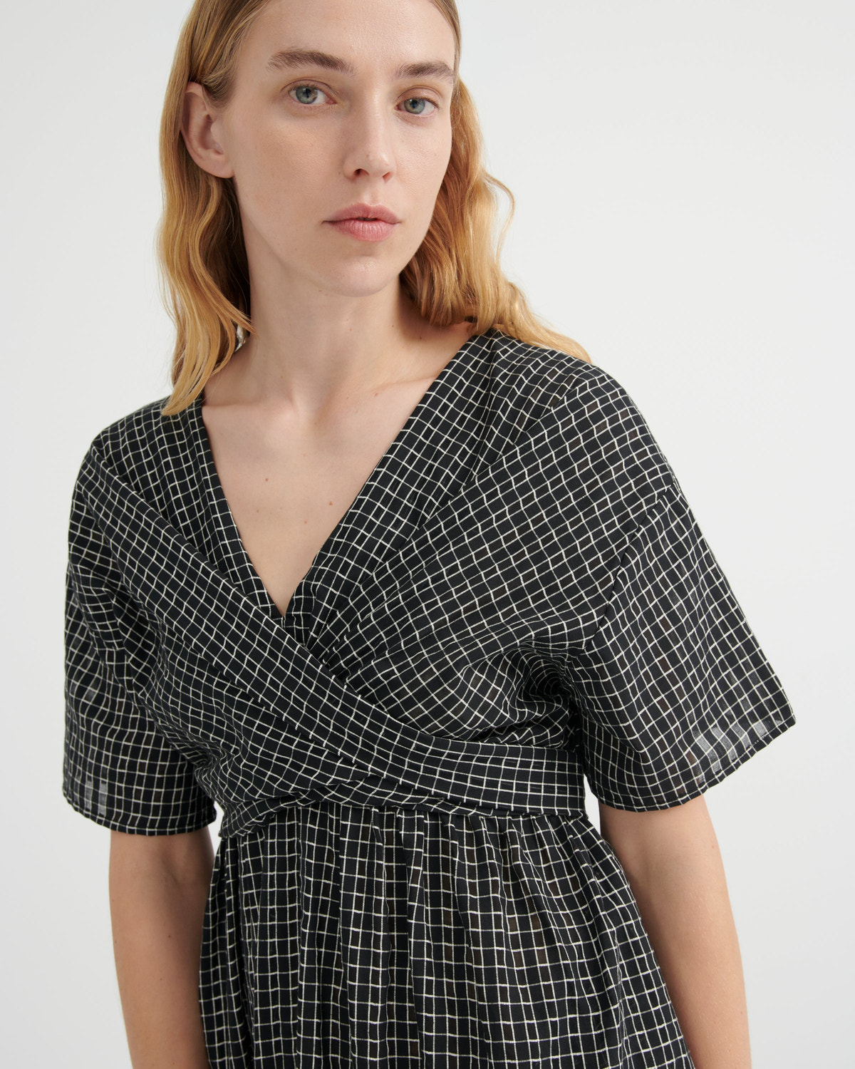 A woman wearing a black and white grid-patterned wrap dress with short puff sleeves and a V-neckline, shown from the chest up against a plain light gray background.