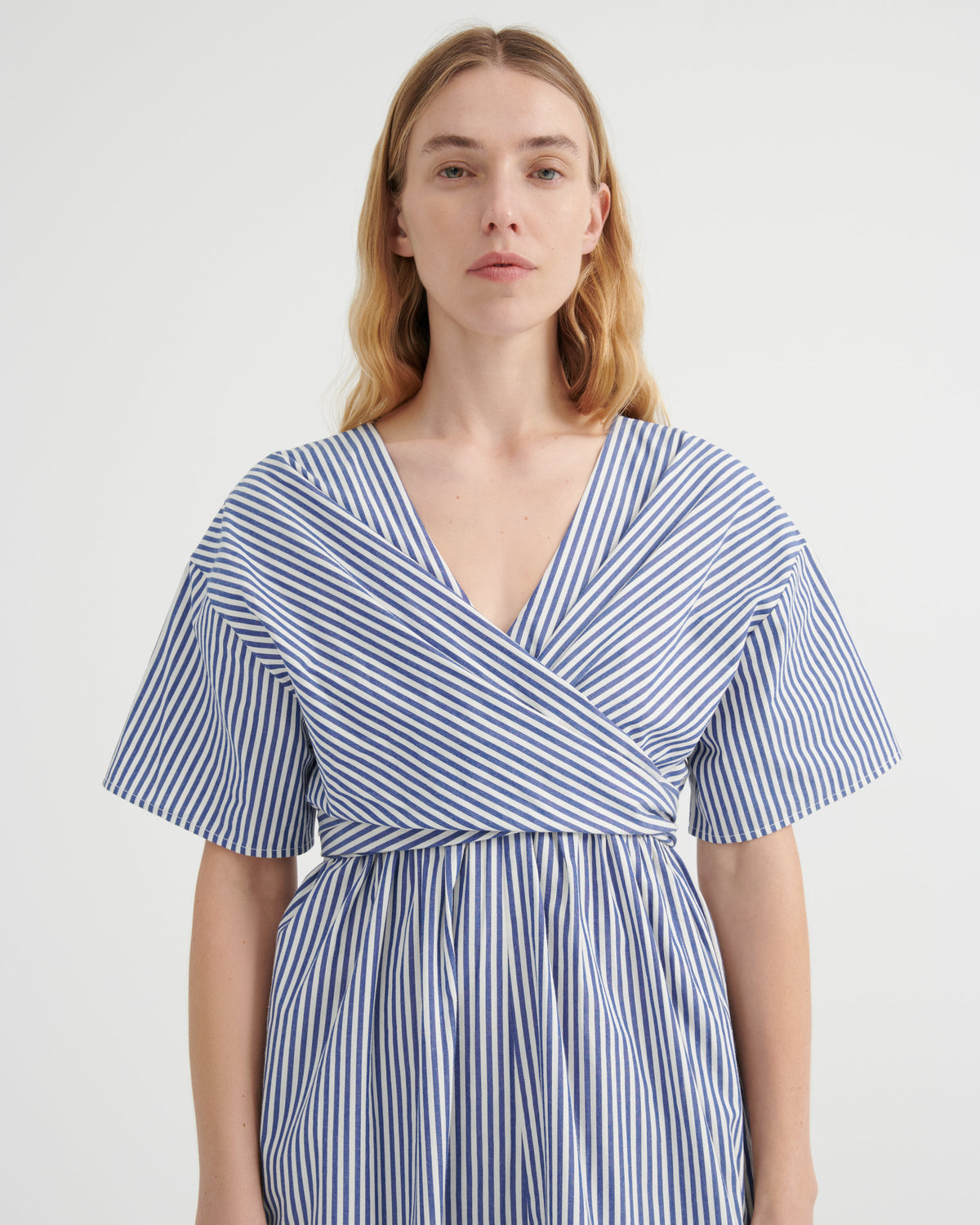 A woman modeling a blue and white vertically striped wrap dress with short flutter sleeves and a V-neckline, photographed against a plain light gray studio background.