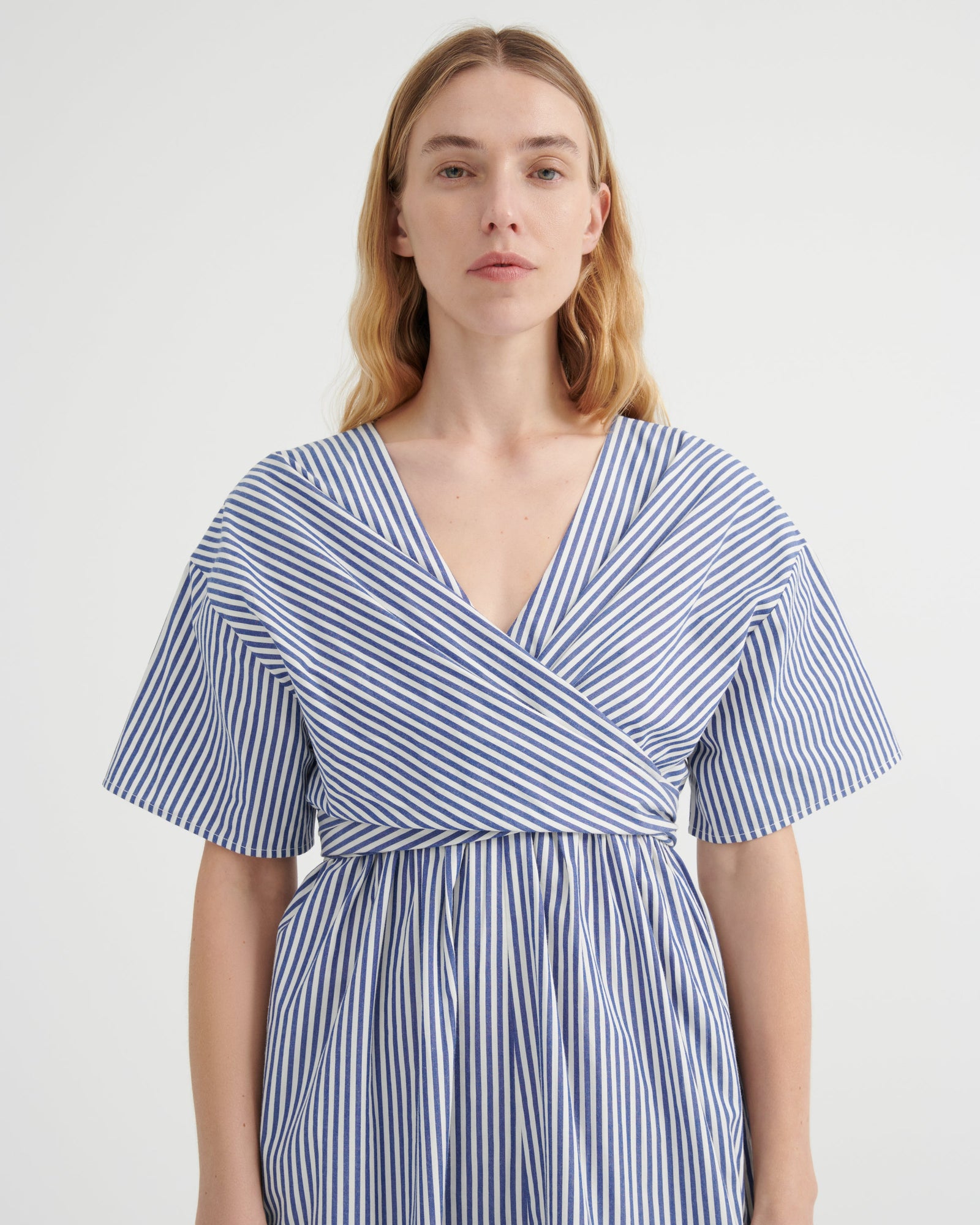 A woman modeling a blue and white vertically striped wrap dress with short flutter sleeves and a V-neckline, photographed against a plain light gray studio background.