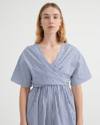 A woman modeling a blue and white vertically striped wrap dress with short flutter sleeves and a V-neckline, photographed against a plain light gray studio background.