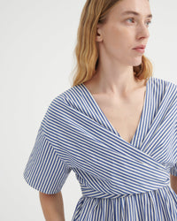 A close-up of a woman wearing a blue and white vertically striped wrap dress with short puff sleeves and a V-neckline, photographed against a clean white studio background.