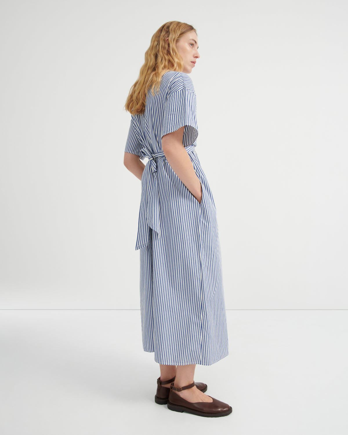 A woman modeling a blue and white vertically striped midi dress with short flutter sleeves, a tie waist, and wide legs, paired with dark brown flat shoes against a plain white studio background.