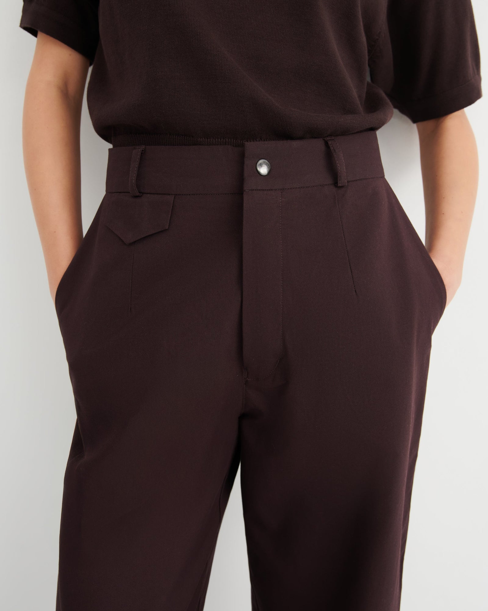 A close-up of a person wearing dark brown tailored trousers and a dark brown short-sleeve top.