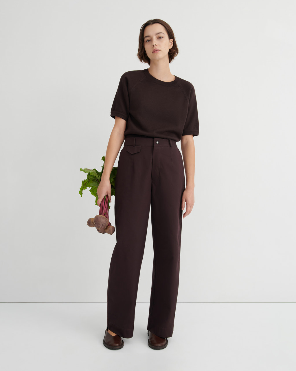 A model stands against a light gray studio background wearing a dark brown short-sleeve crewneck top and matching dark brown wide-leg trousers. She is holding a bunch of beetroots.