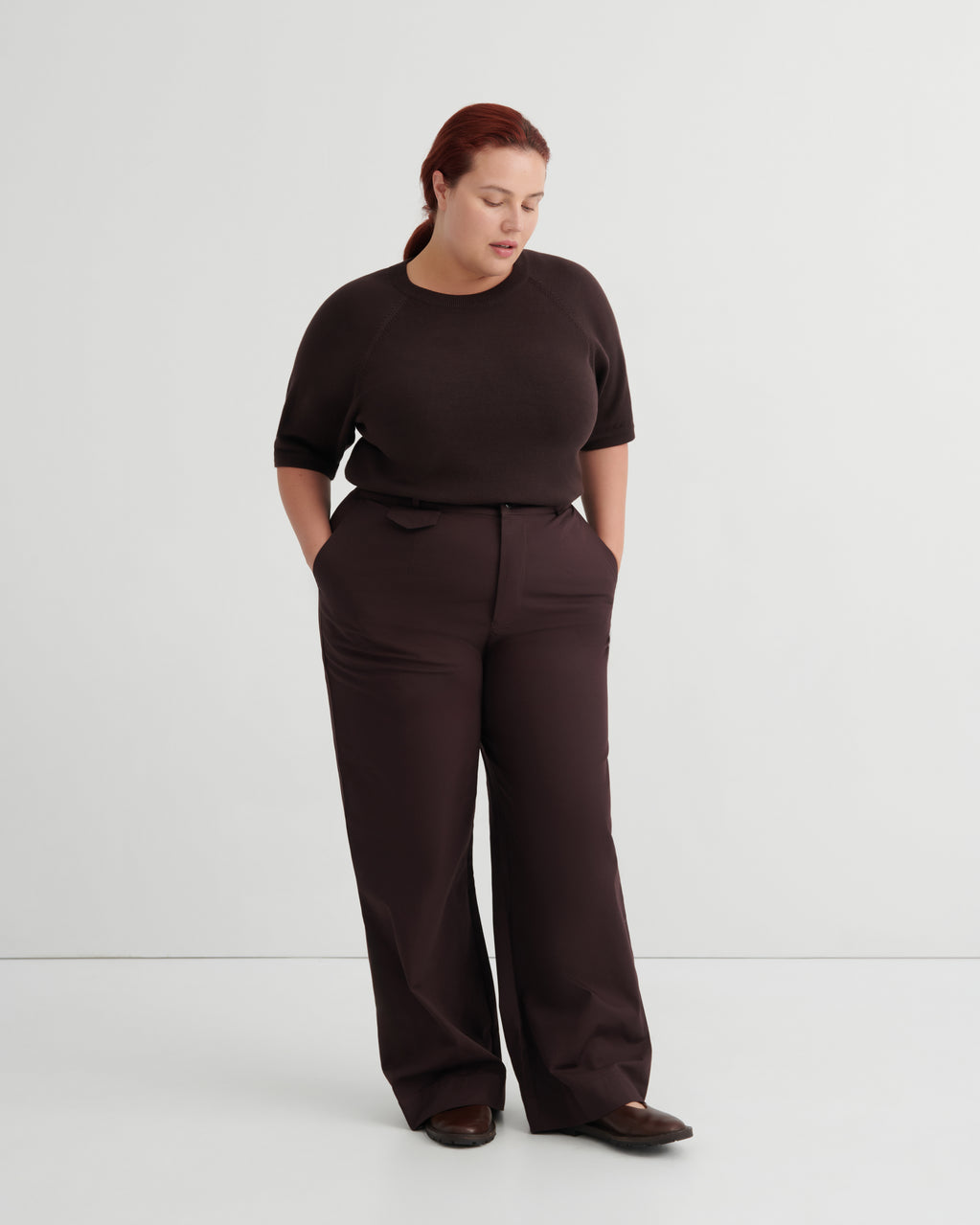 A woman modeling a dark brown short-sleeve top and matching wide-leg trousers against a plain light gray studio background.