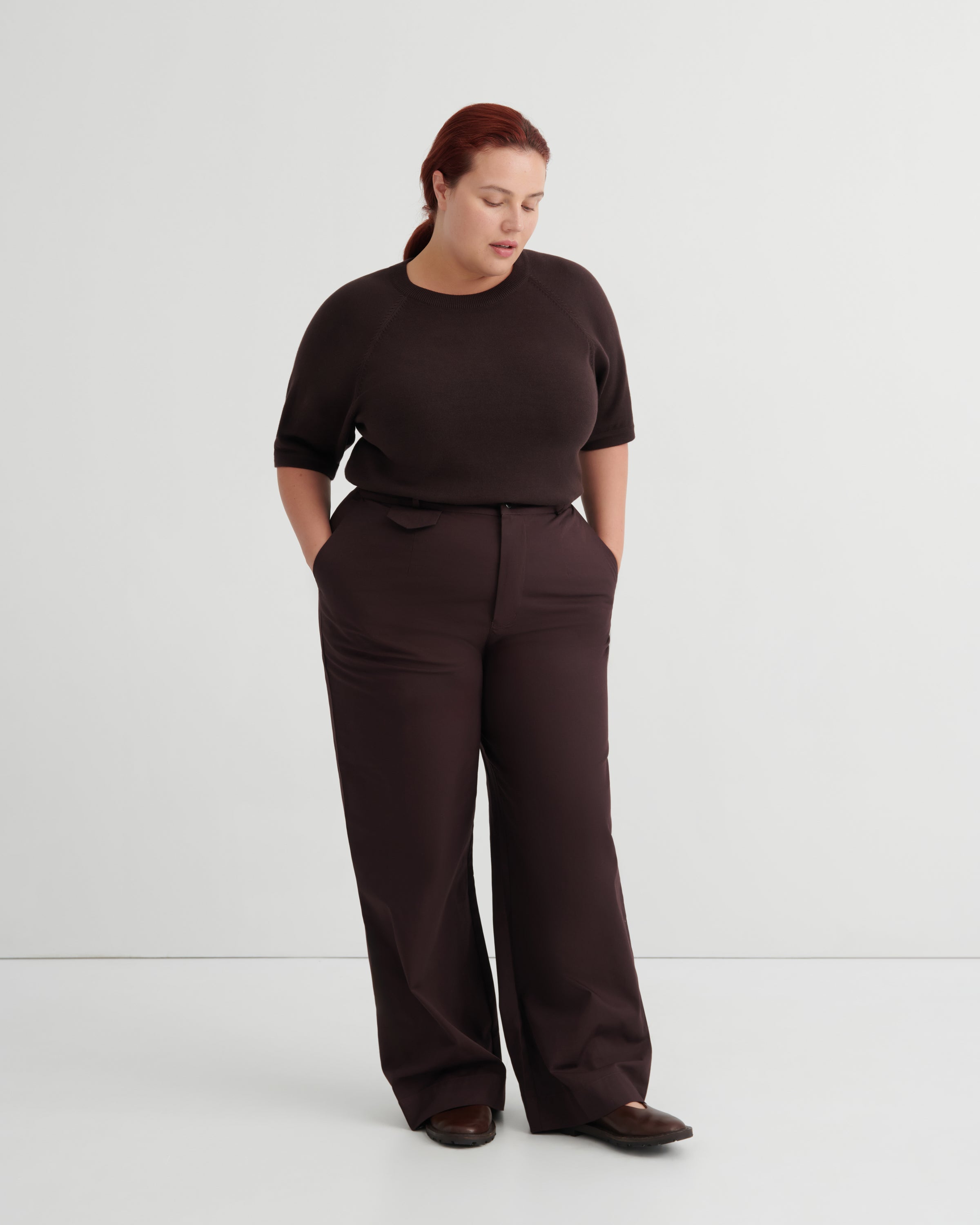 A woman modeling a dark brown short-sleeve top and matching wide-leg trousers against a plain light gray studio background.