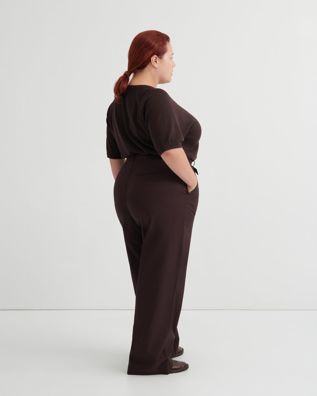 A woman with red hair tied back is shown from the back wearing a dark brown short-sleeve top and matching dark brown wide-leg trousers, standing against a plain light gray studio background.