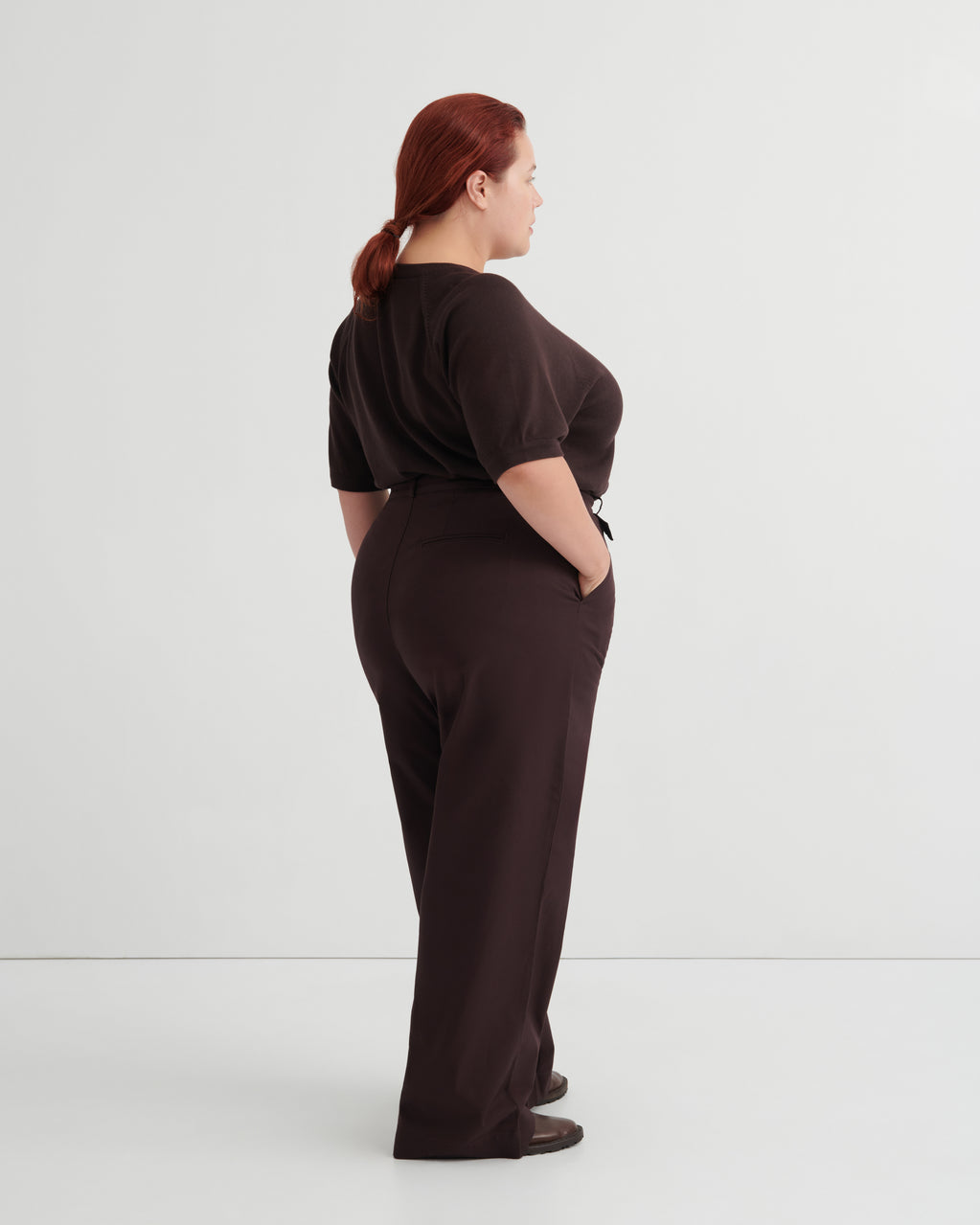 A woman with red hair tied back is shown from the back wearing a dark brown short-sleeve top and matching dark brown wide-leg trousers, standing against a plain light gray studio background.