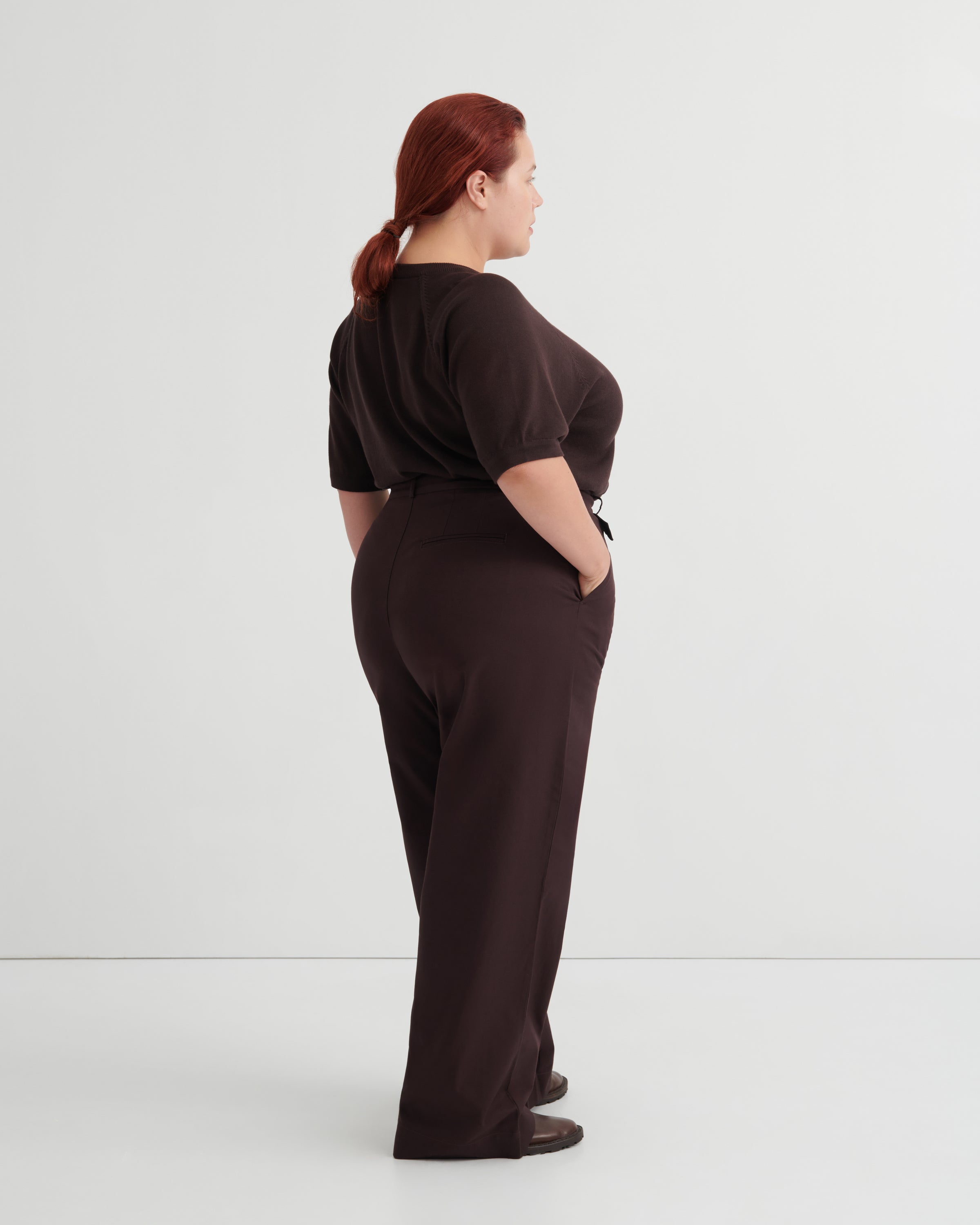 A woman with red hair tied back is shown from the back wearing a dark brown short-sleeve top and matching dark brown wide-leg trousers, standing against a plain light gray studio background.