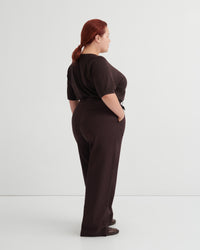 A woman with red hair tied back is shown from the back wearing a dark brown short-sleeve top and matching dark brown wide-leg trousers, standing against a plain light gray studio background.