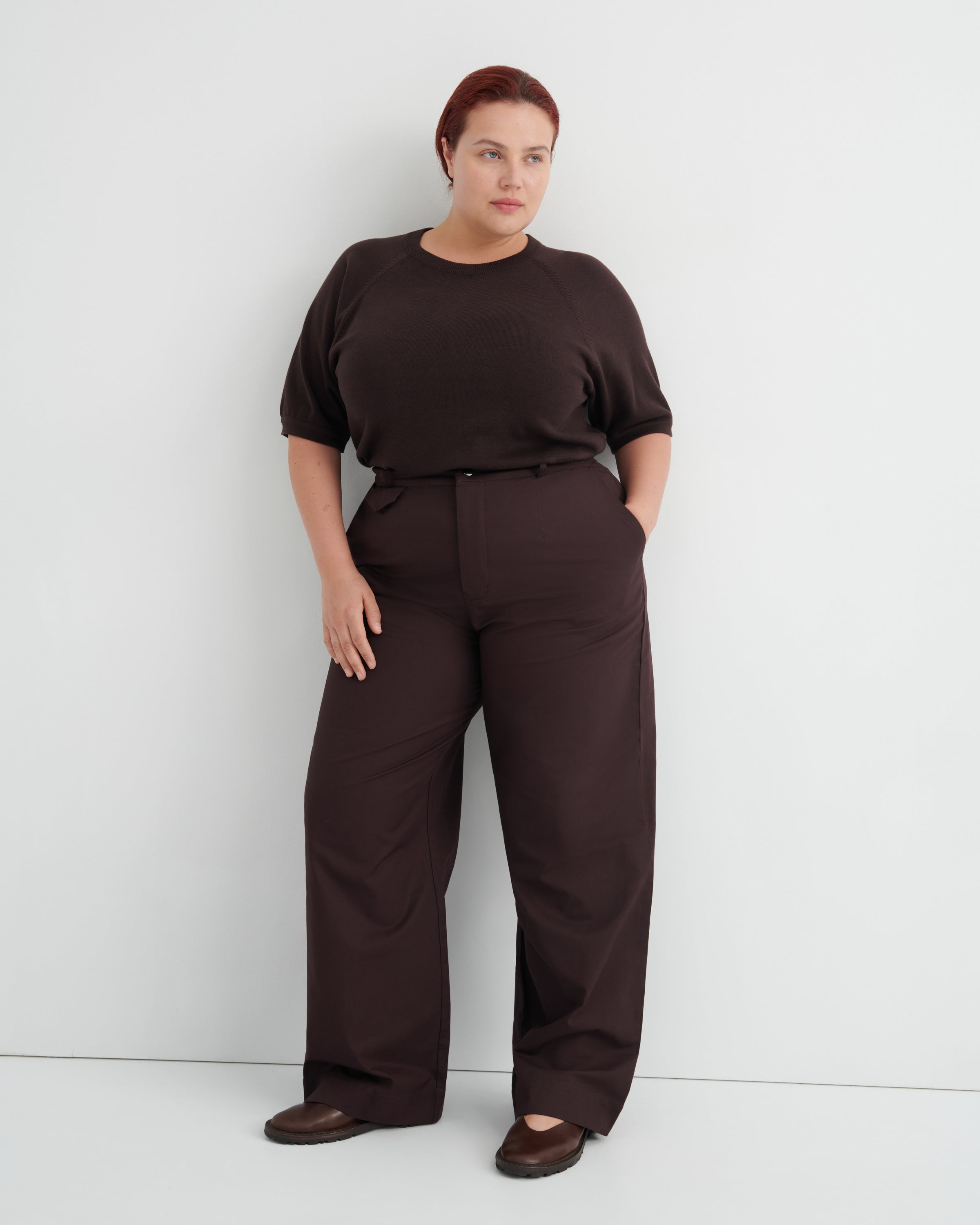 A woman modeling a dark brown short-sleeve top and matching wide-leg trousers with a thin black belt, standing against a light gray studio wall.