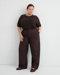 A woman modeling a dark brown short-sleeve top and matching wide-leg trousers with a thin black belt, standing against a light gray studio wall.