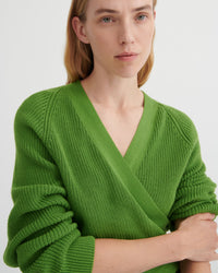 A close-up of a woman wearing a bright green ribbed knit V-neck wrap sweater, showing the texture and fit against a plain light background.