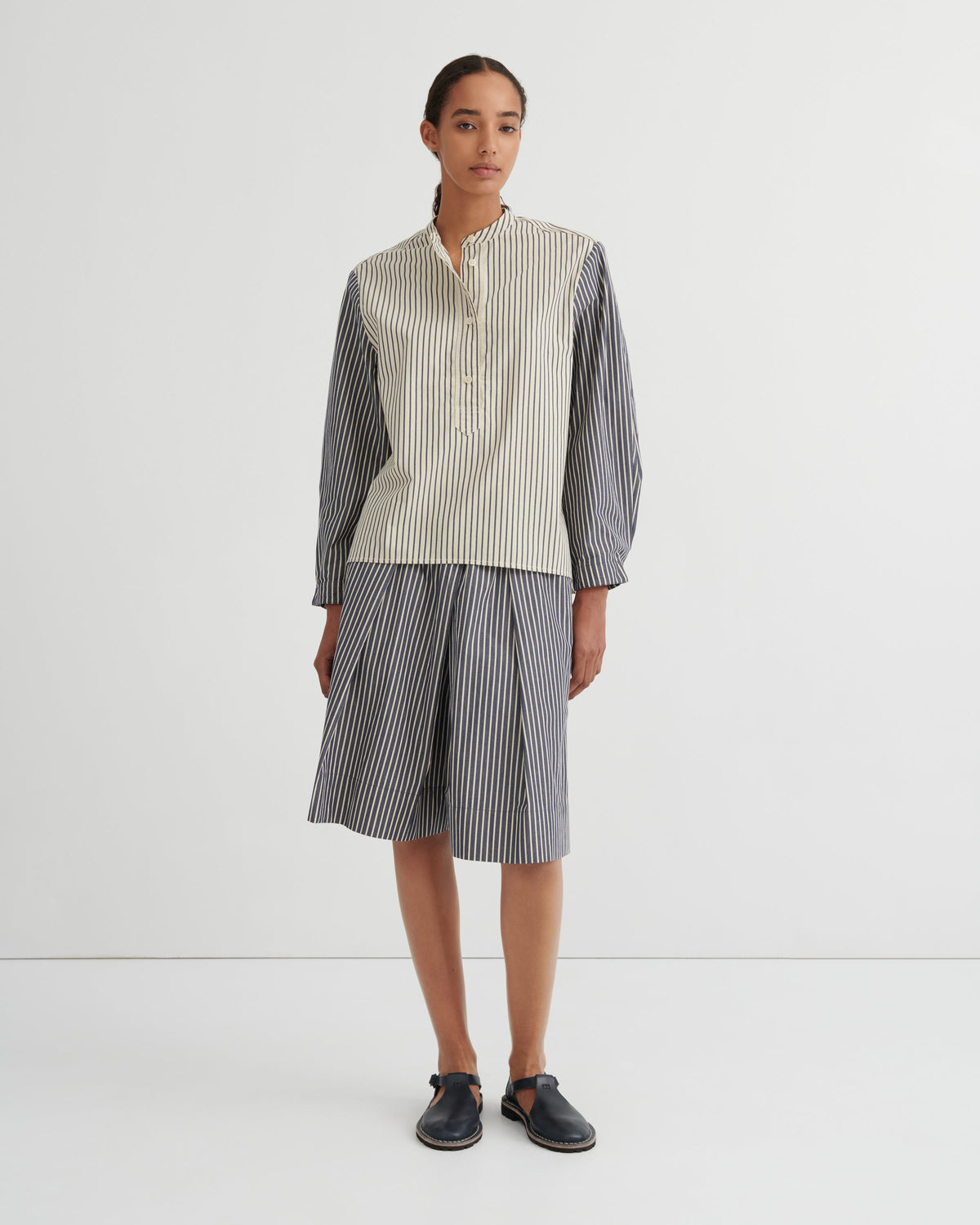 A model stands against a plain white studio background wearing a matching striped two-piece set consisting of a button up shirt and pleated shorts. The shirt has a cream and white vertical stripe pattern on the front and solid dark gray sleeves, while the shorts are entirely dark gray with thin white vertical stripes.