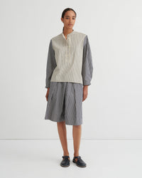 A model stands against a plain white studio background wearing a matching striped two-piece set consisting of a button up shirt and pleated shorts. The shirt has a cream and white vertical stripe pattern on the front and solid dark gray sleeves, while the shorts are entirely dark gray with thin white vertical stripes.