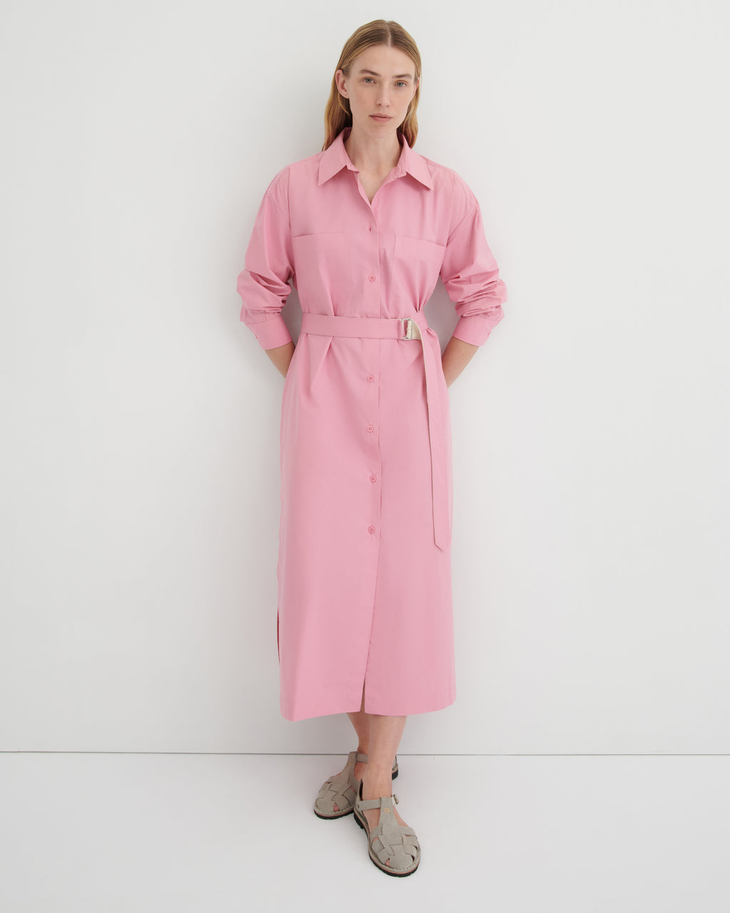 A women wearing a button up pink dress with the sleeves rolled up and the belt cinching the waist.