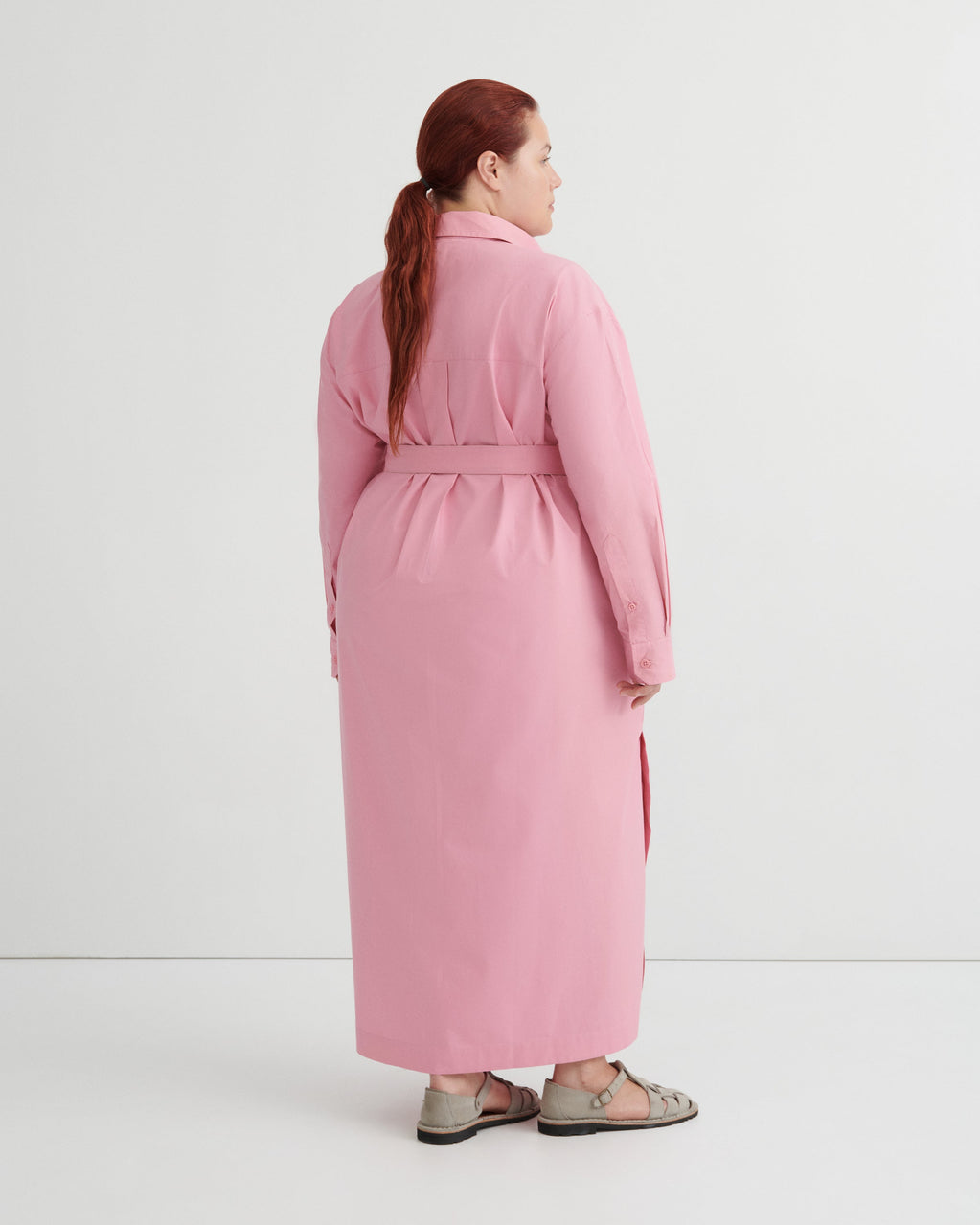 A woman with red hair tied back in a ponytail is shown from the back wearing a long pink belted coat or dress over a light gray top and gray sandals, photographed against a clean white studio background.