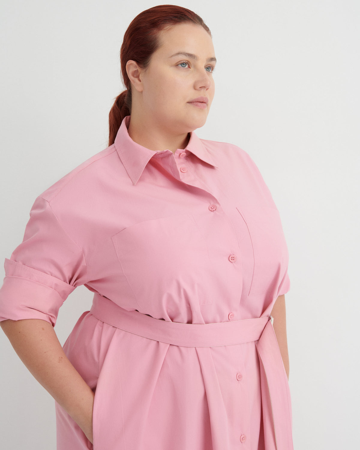 A woman wearing a light pink collared shirt dress with rolled sleeves and a matching waist tie, photographed against a plain light gray studio background.