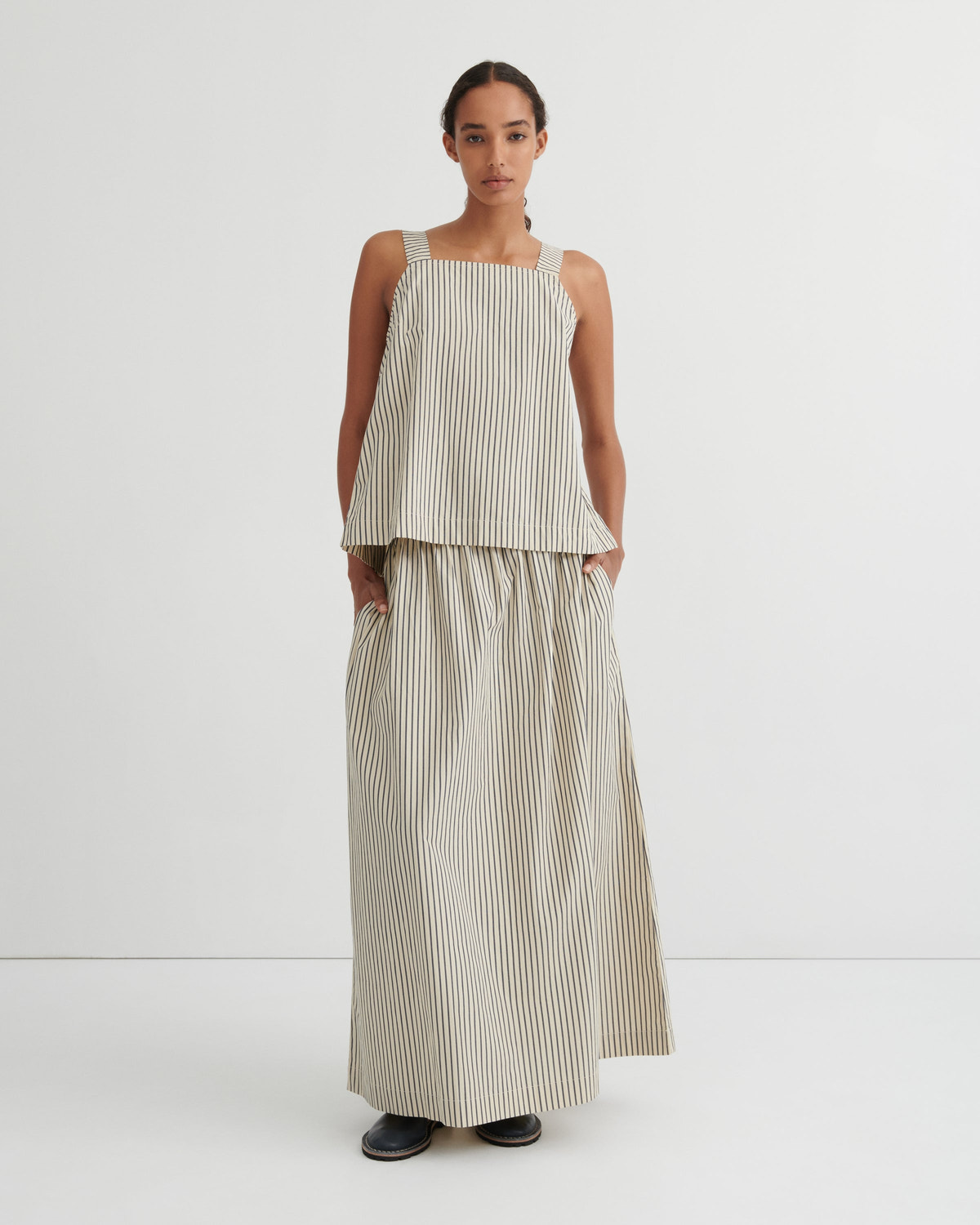 A model stands against a plain white studio background wearing a sleeveless beige and white vertically striped two-piece set consisting of a cropped top and matching wide-leg skirt.