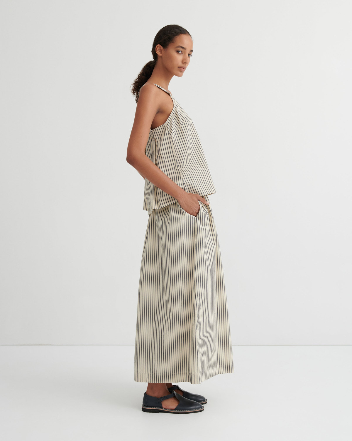 A model is shown in profile wearing a matching beige and white vertically striped two-piece set consisting of a halter crop top and wide-leg trousers, paired with dark navy flat shoes, against a plain white studio background.