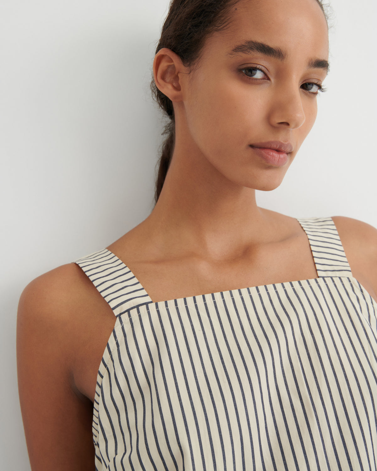 A close-up of a model wearing a sleeveless top with thin vertical navy stripes on a cream background, set against a plain light gray wall.