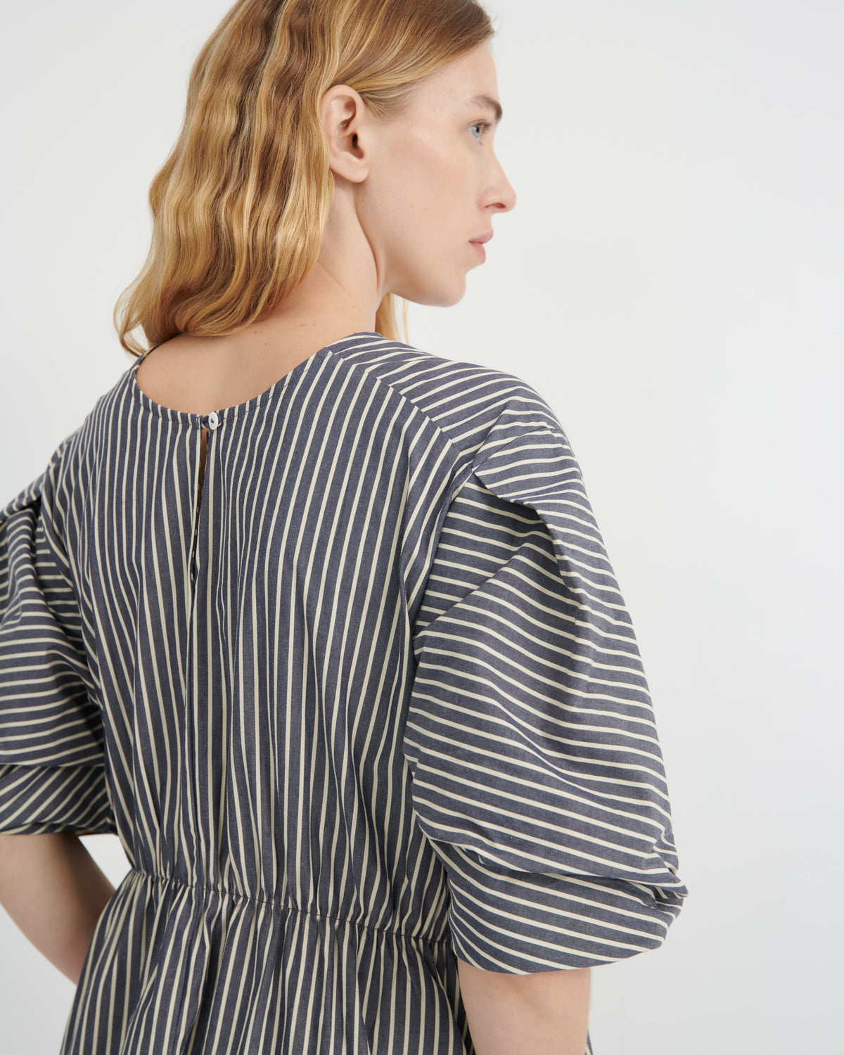 A woman shown from the back wearing a dark gray and white vertically striped dress with puff sleeves and a gathered waist, photographed against a clean white studio background.