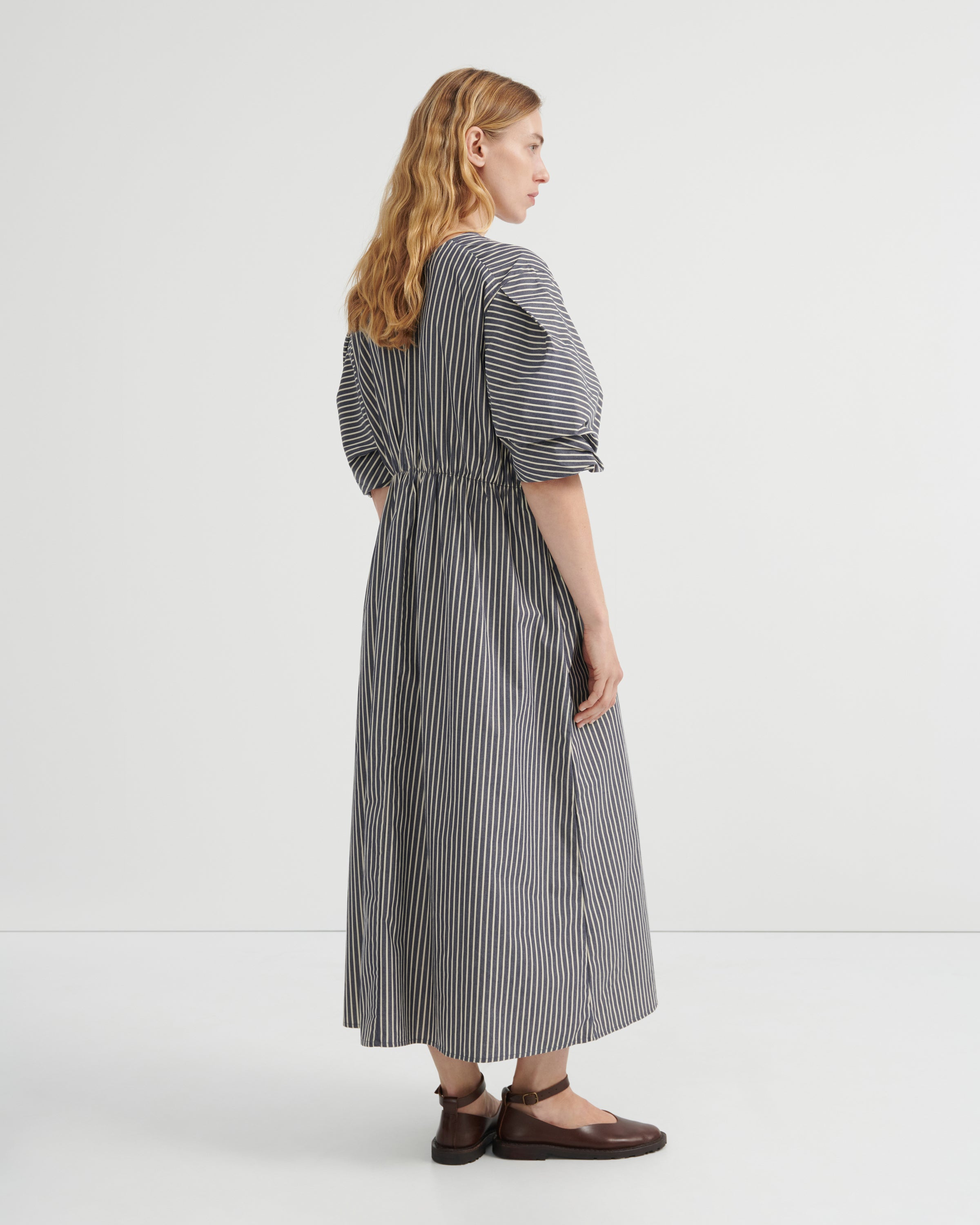 A woman modeling a long-sleeved midi dress with vertical grey and white stripes, puff sleeves, and a gathered waist, shown from a side-back angle against a plain white studio background.