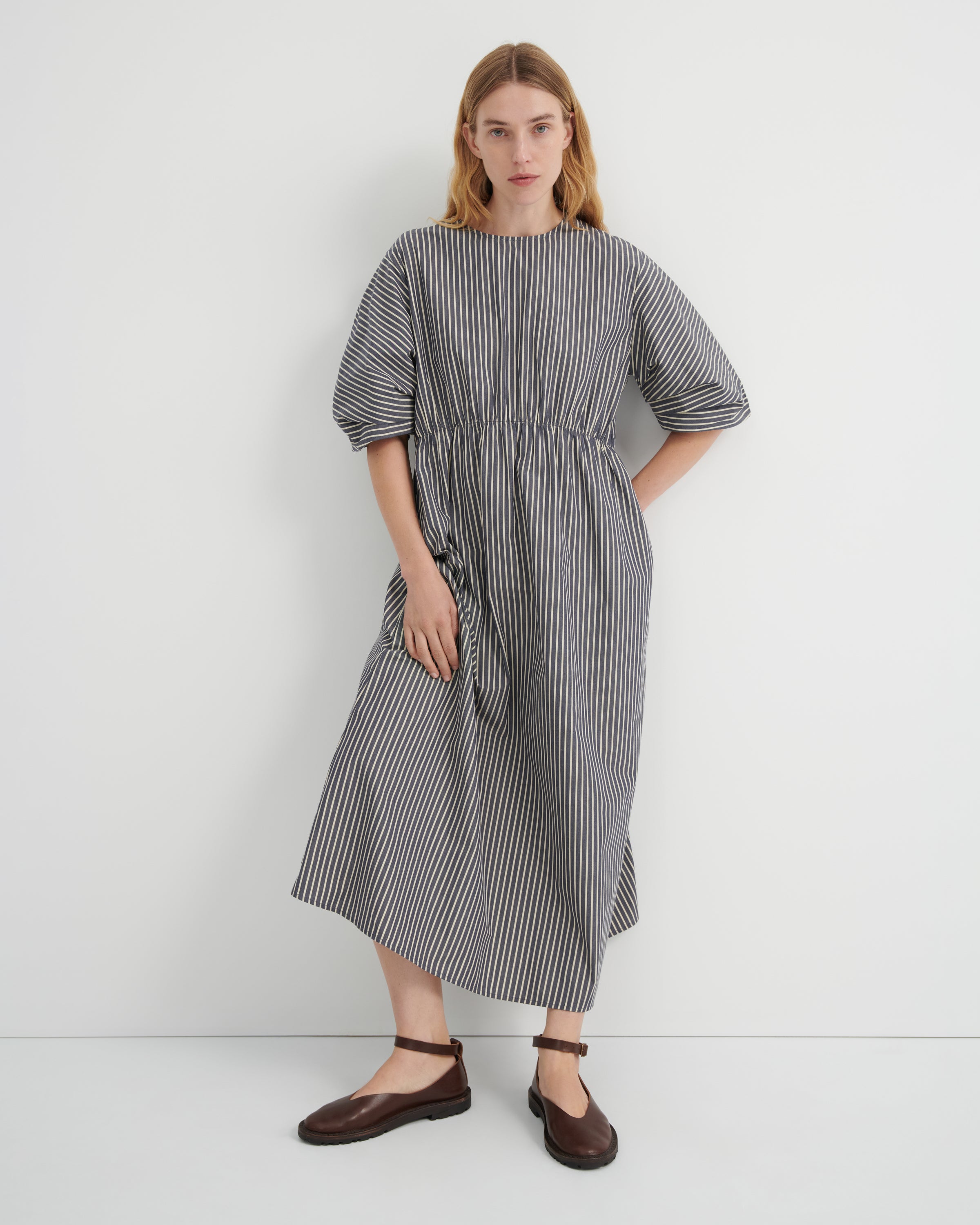 A model wearing a striped grey and white midi dress with a gathered waist and puff sleeves against a grey background.