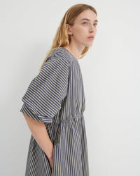 A woman wearing a loose-fitting, short-sleeved dress with vertical gray and white stripes, shown from a side profile against a plain light gray background.