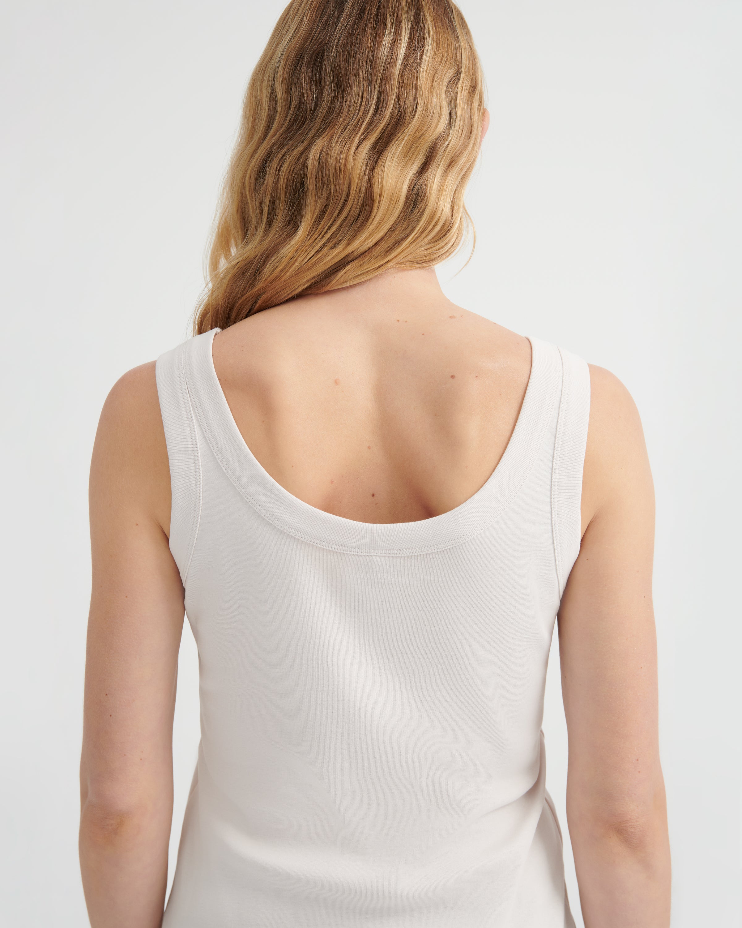 A woman viewed from the back wearing a light off-white scoop-neck tank top.
