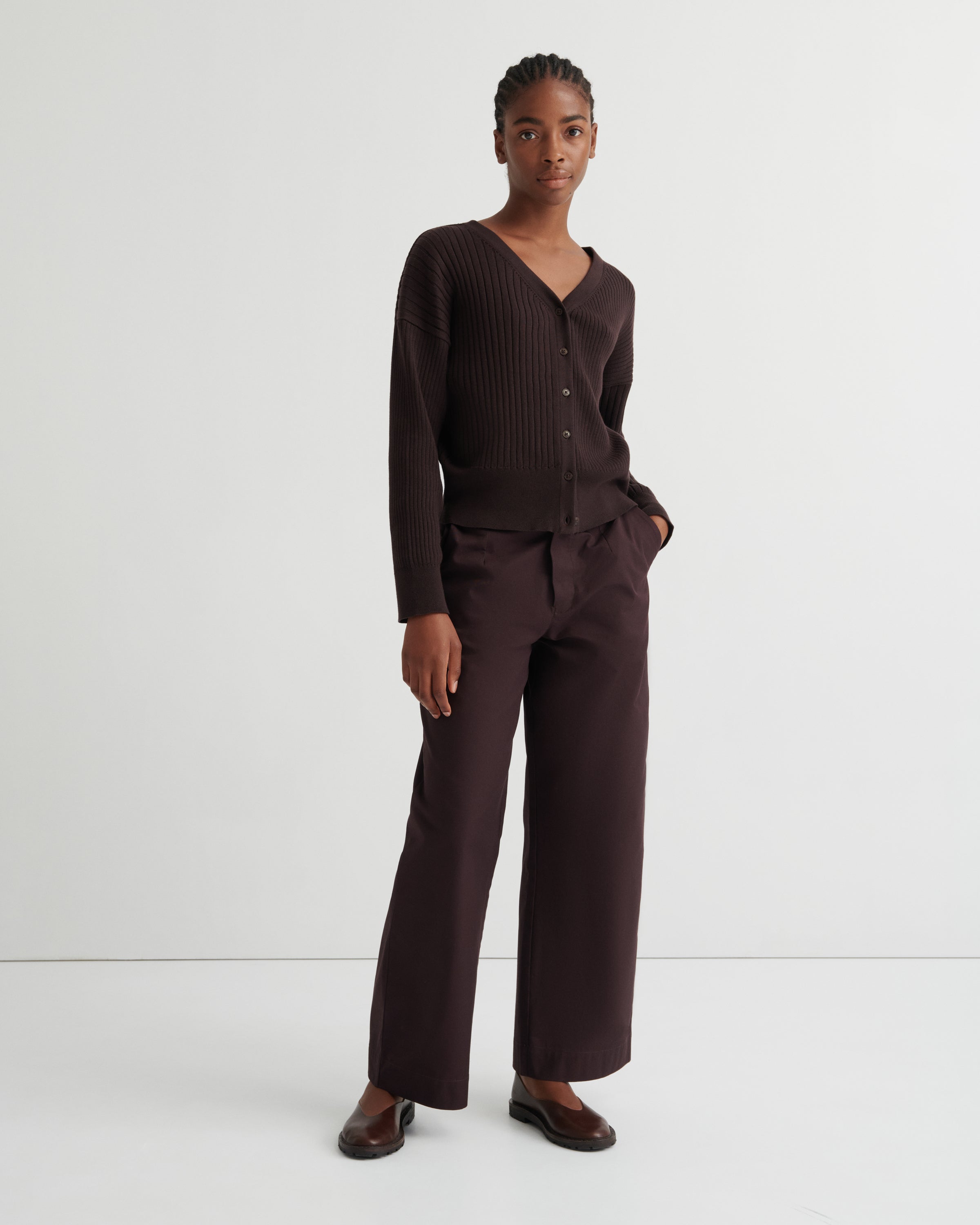 A model stands against a plain light gray studio background wearing a dark brown ribbed knit cardigan with a V-neck and buttons, matching wide-leg trousers, and dark brown loafers.