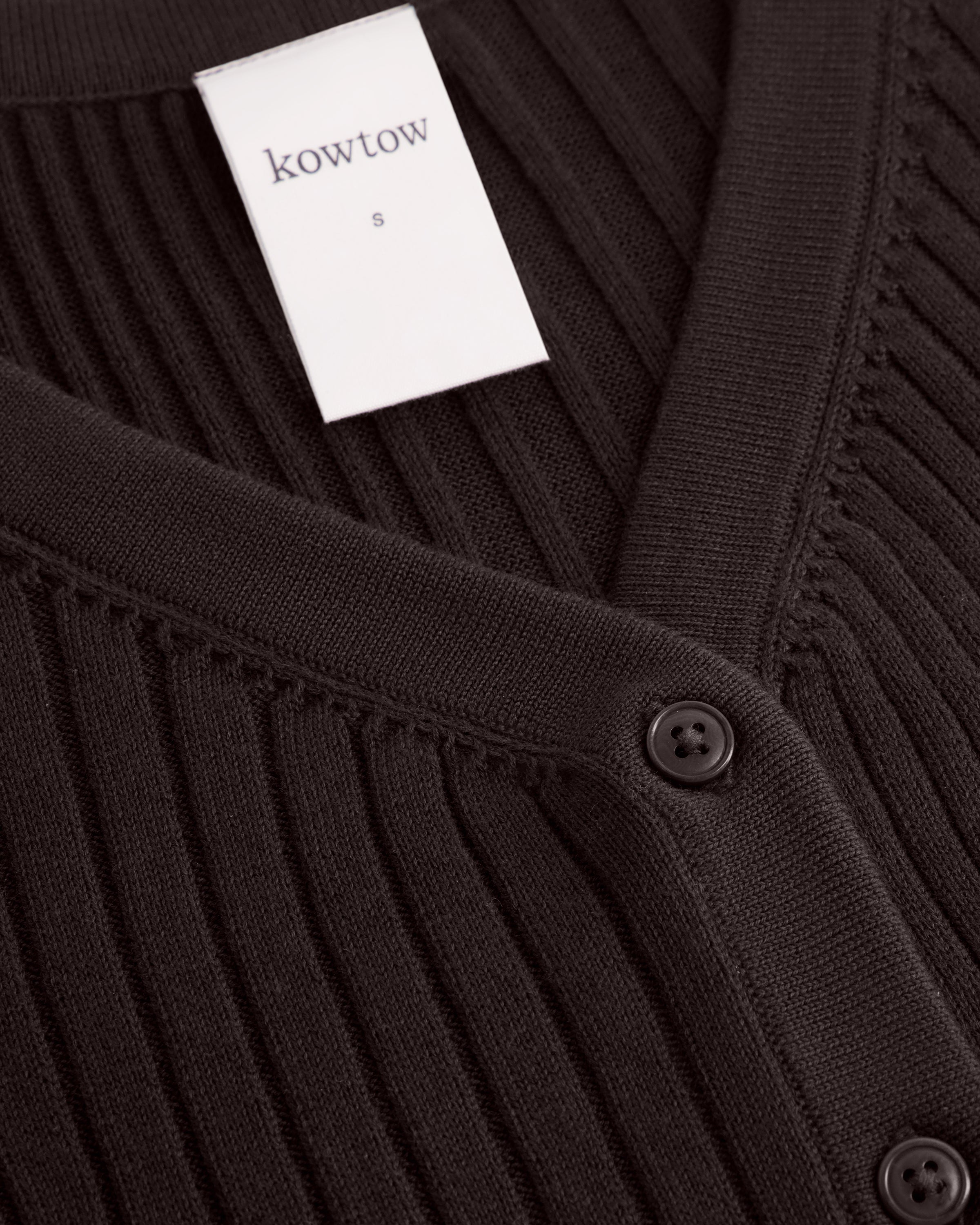 Close-up of a dark brown ribbed knit cardigan sweater with a white Kowtow brand tag and dark buttons.