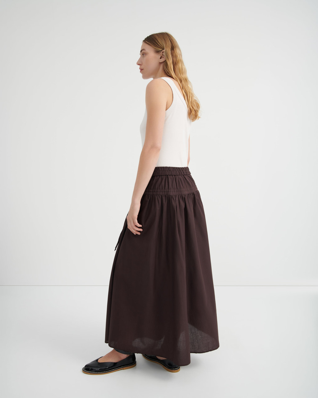A woman shown from the side profile wearing a white sleeveless top and a long, dark brown gathered skirt.