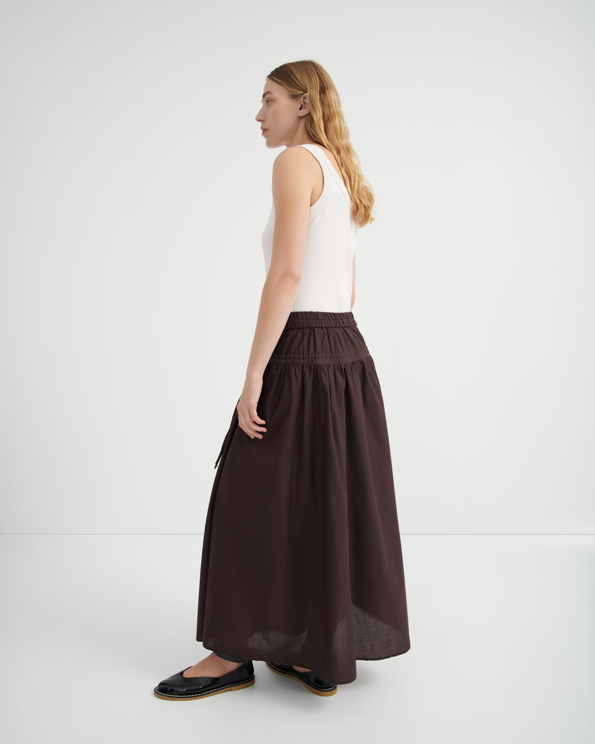 A woman shown from the side profile wearing a white sleeveless top and a long, dark brown gathered skirt.