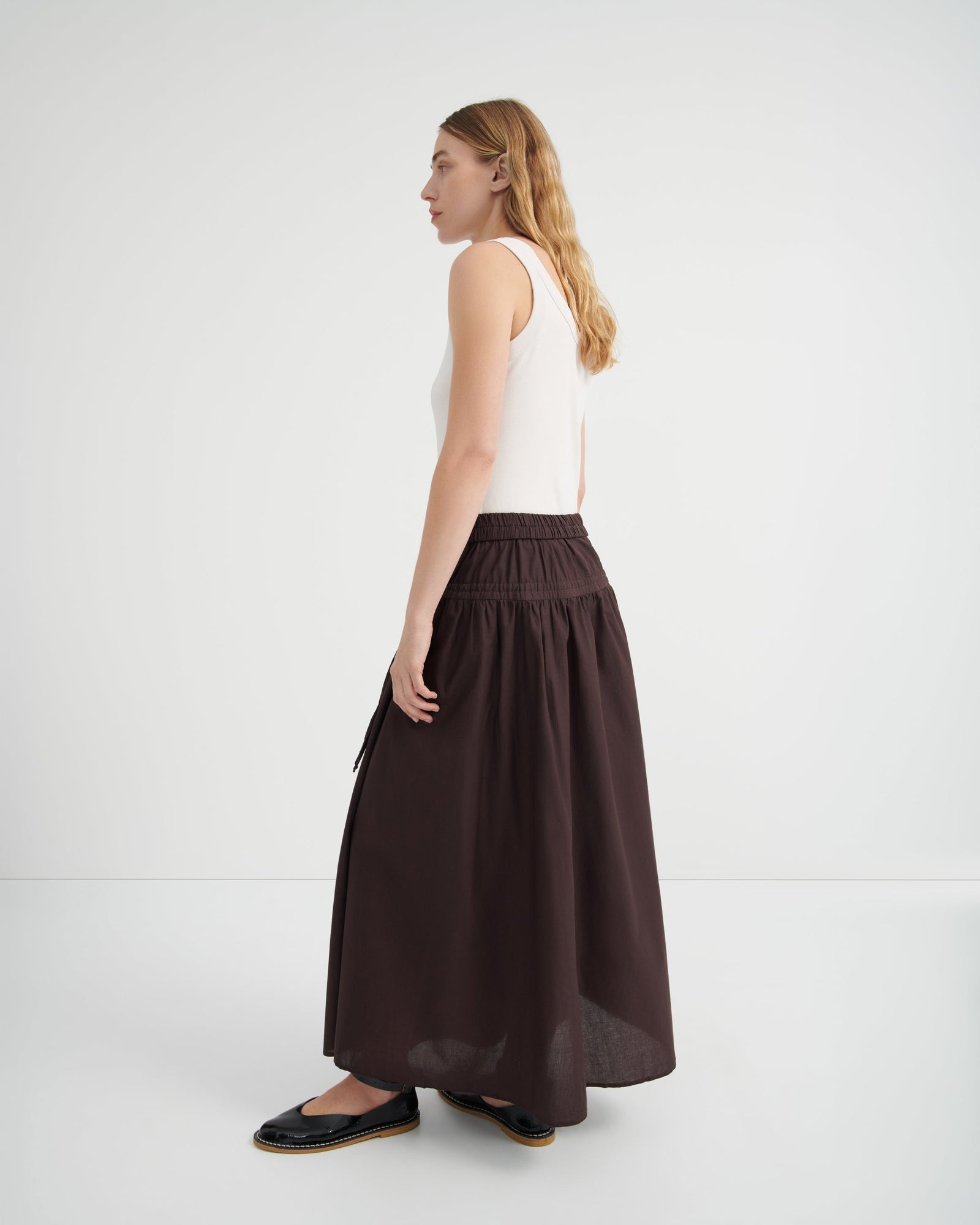 A woman shown from the side profile wearing a white sleeveless top and a long, dark brown gathered skirt.