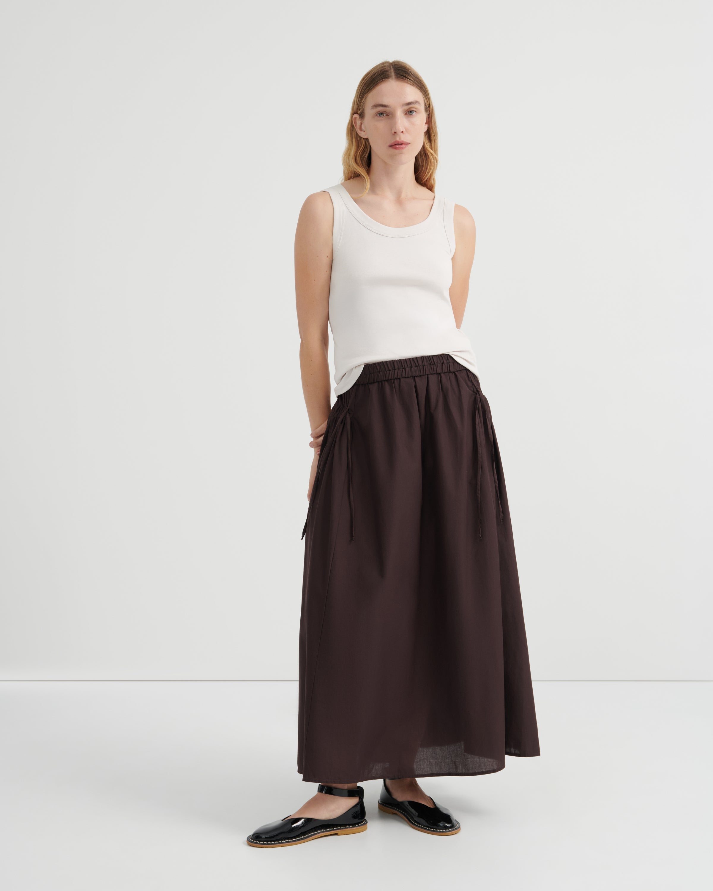 A woman modeling a dark brown high-waisted ruched maxi skirt paired with a white sleeveless top and black flat shoes.