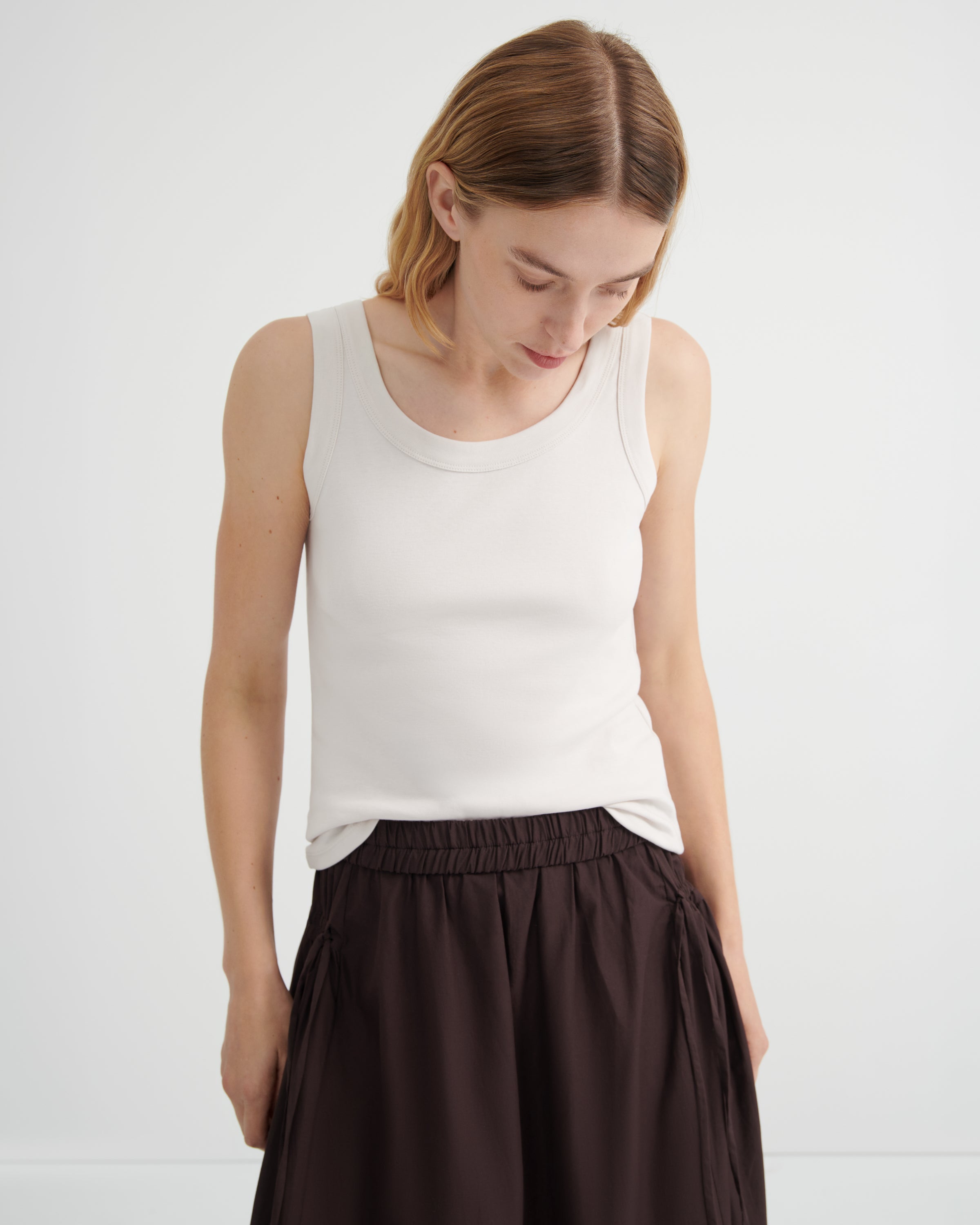 A woman wearing a fitted white ribbed tank top and dark brown pleated skirt, looking down against a plain light gray studio background.