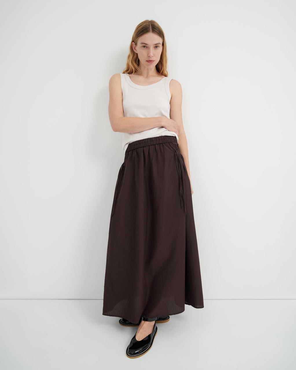 A model wearing a sleeveless white top and a long dark brown skirt with a tie waist, paired with black flats.