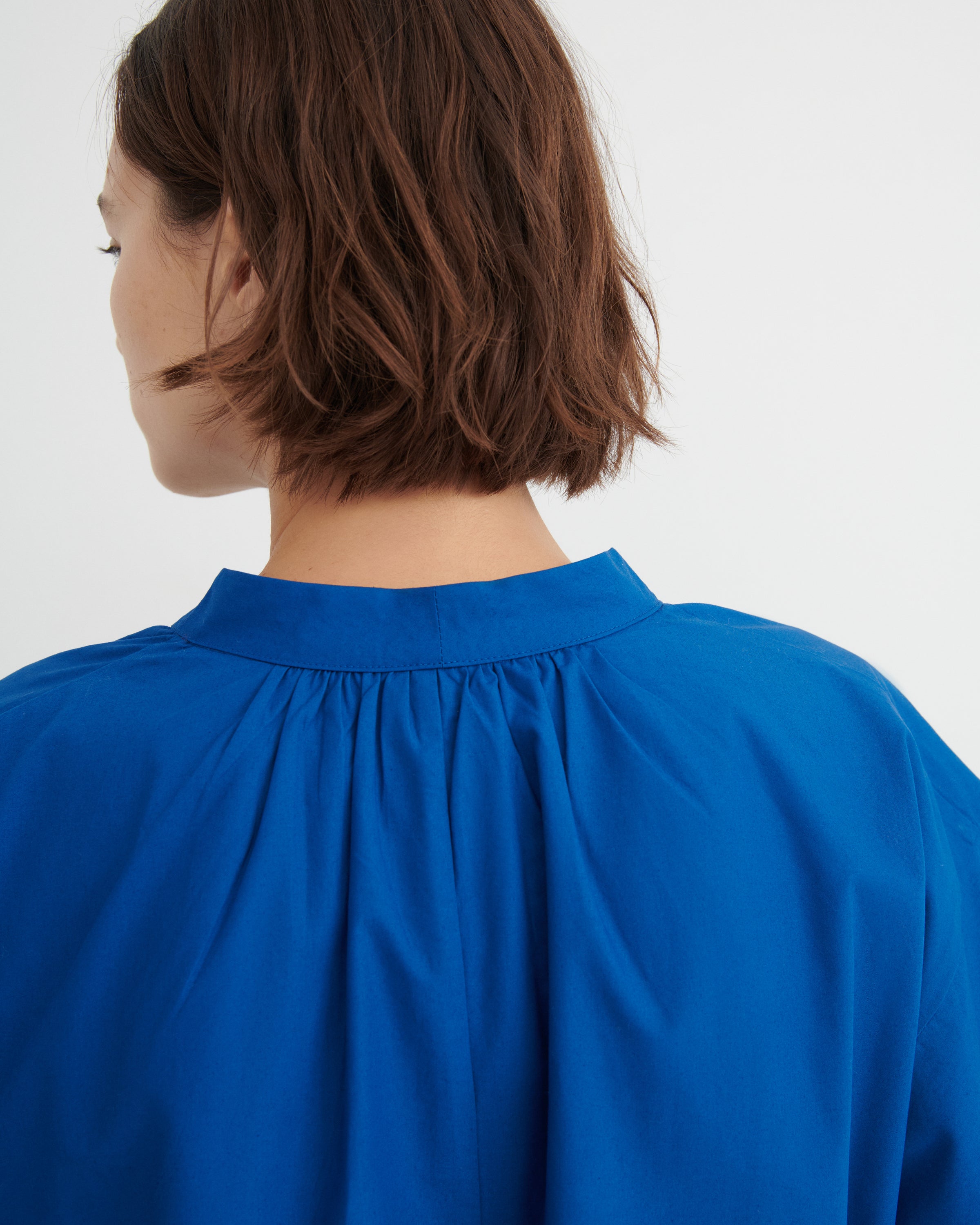 A close-up back view of a person with short brown hair wearing a bright royal blue blouse with a gathered, pleated neckline against a plain light background.