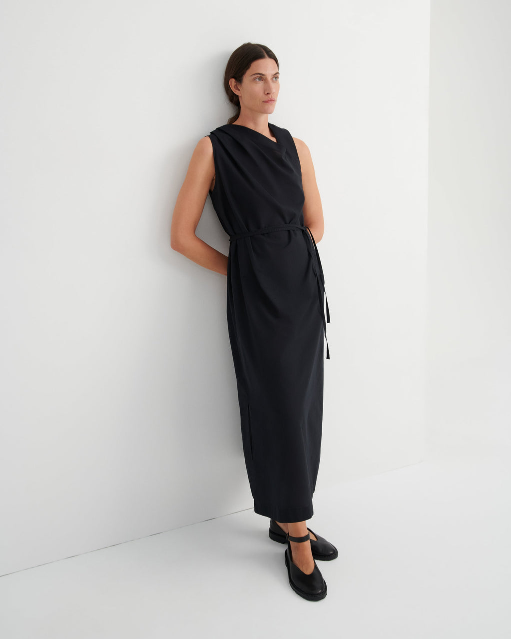 A woman wearing a sleeveless black midi dress with a tie waist and black Mary Jane shoes, standing against a clean white wall.