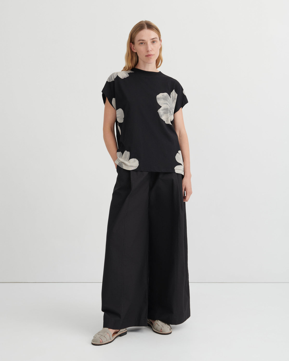 A model stands against a light gray studio background wearing a black short-sleeve top with large white floral applique details, paired with wide-leg black trousers and gray slip-on shoes.