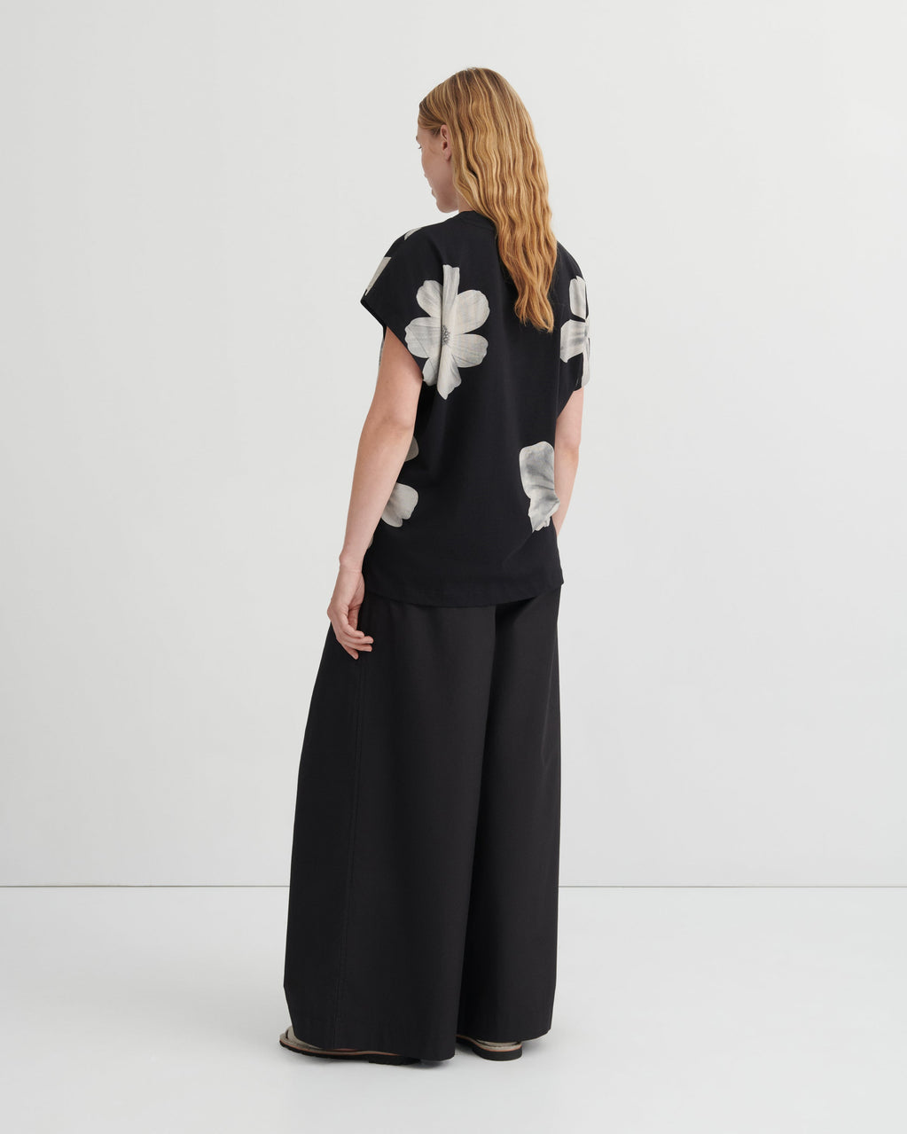 A woman stands with her back to the camera wearing a black short-sleeve top with large white floral applique and black wide-leg pants, photographed in a clean studio setting.
