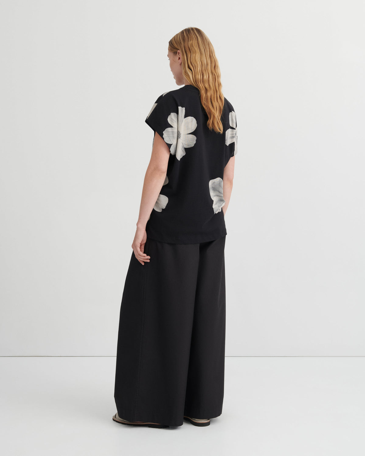 A woman stands with her back to the camera wearing a black short-sleeve top with large white floral applique and black wide-leg pants, photographed in a clean studio setting.