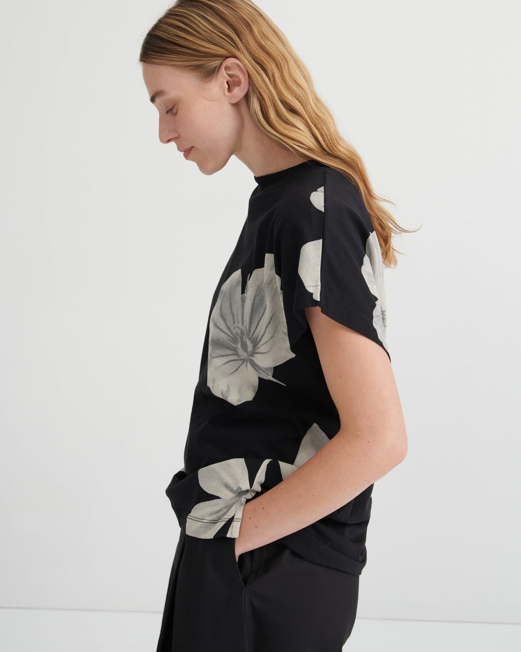 A woman shown in side profile wearing a black short-sleeve top with a large light gray floral print and black pants, photographed against a clean white studio background.